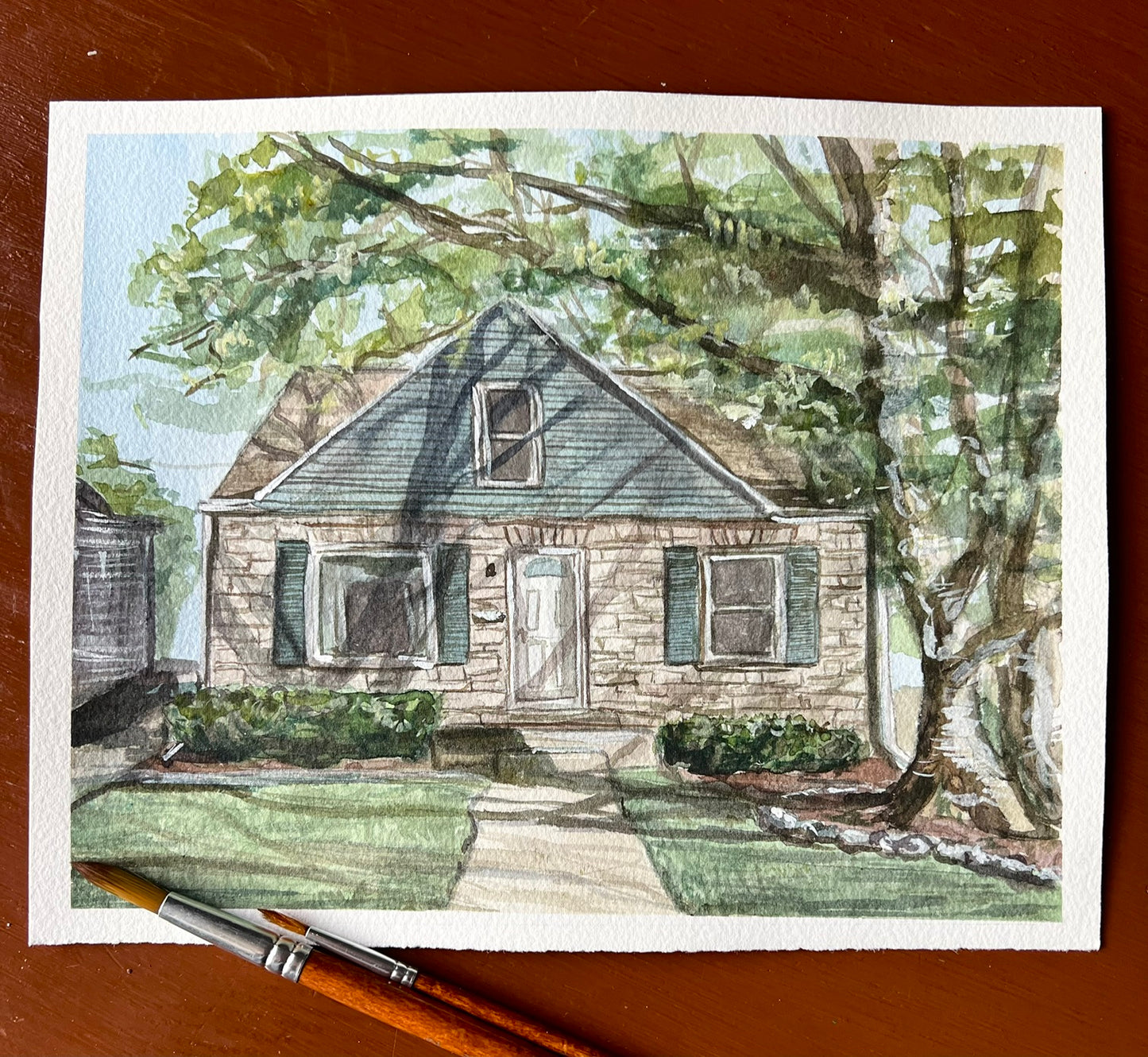 This is a watercolor painting of a house with two paint brushes laying across it 