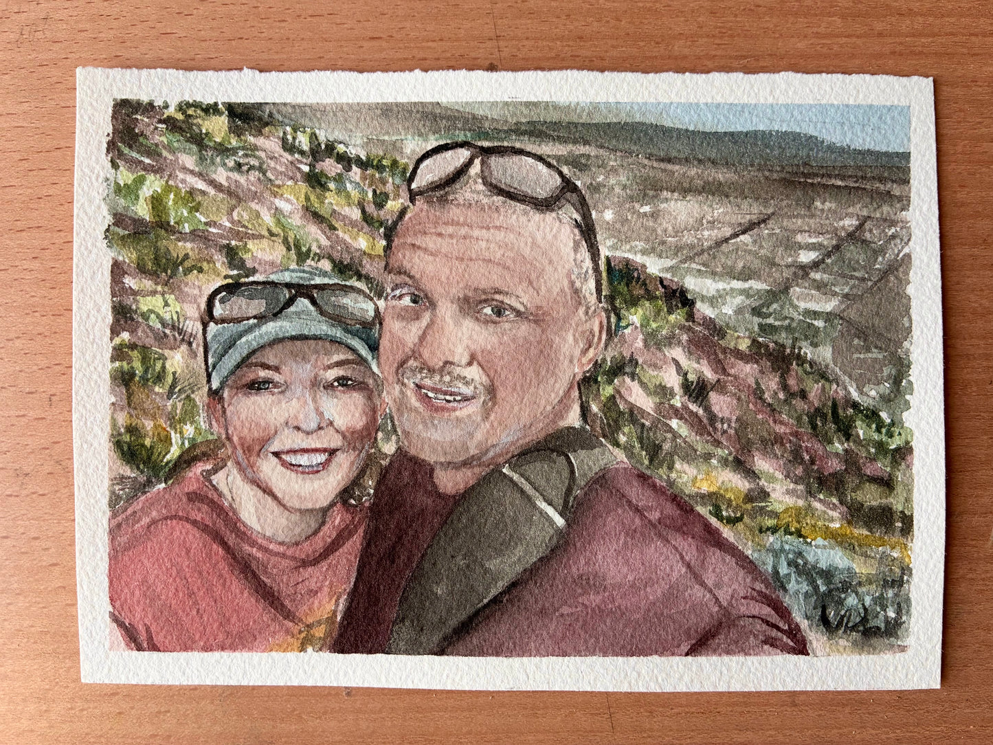 this watercolor painting is two people smiling on a mountain top with a city below them